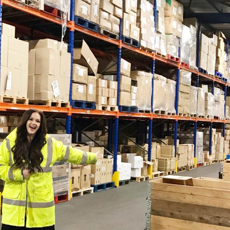 Person in a warehouse wearing a high-visibility jacket with shelves filled with boxes in the background.