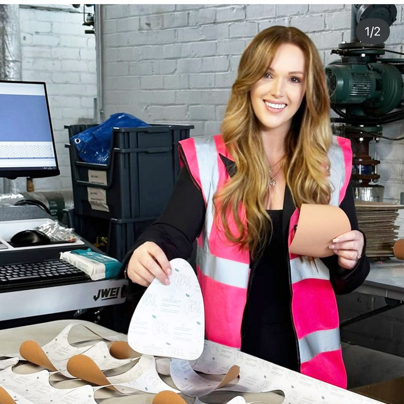 Woman in a workshop holding a shoe insole and a foam block, wearing a pink safety vest.